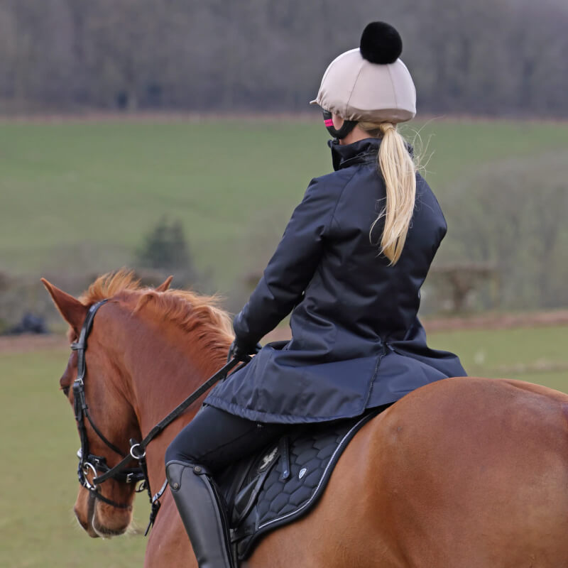 Person in a Shires Aubrion React Mid Length Coat Shadow and helmet riding a brown horse in grass.
