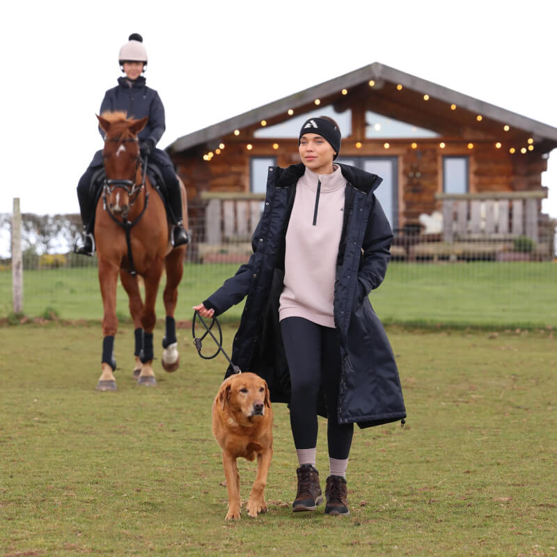 A woman in a Shires Aubrion React Long Coat Shadow walks her dog as someone rides past a cabin.