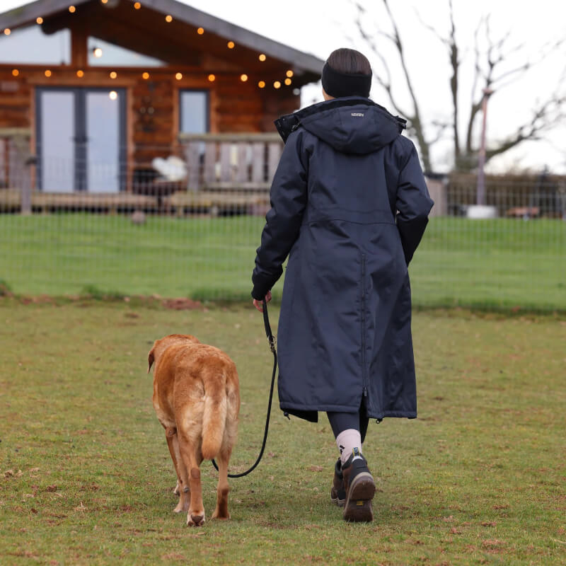 Person in a Shires Aubrion React Long Coat Shadow walks a brown dog near a wooden house.