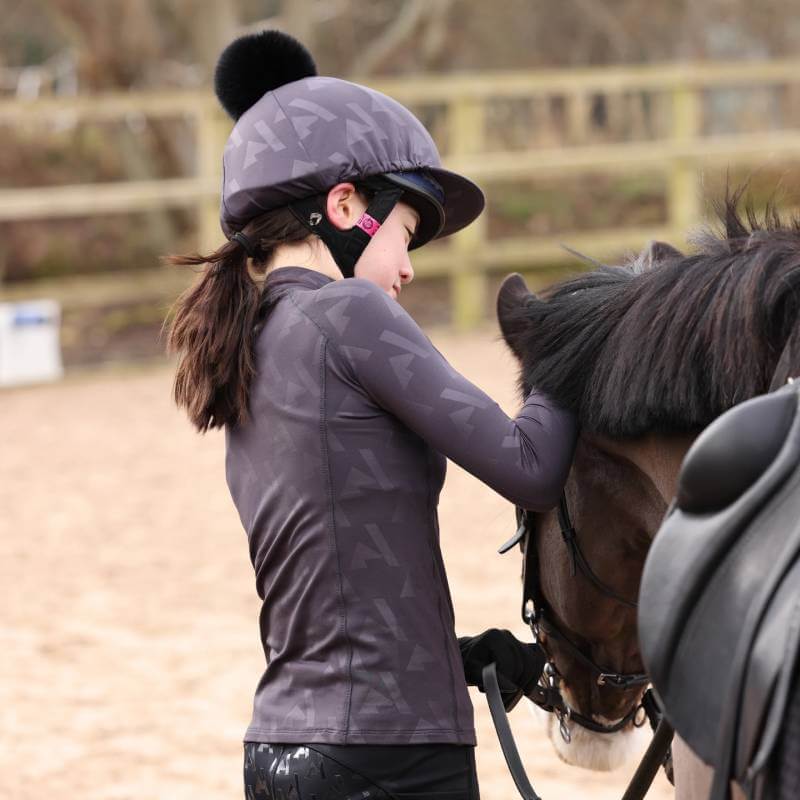 Girl in riding gear and Aubrion React Winter Baselayer YR Shadow pats a dark brown horse outdoors.
