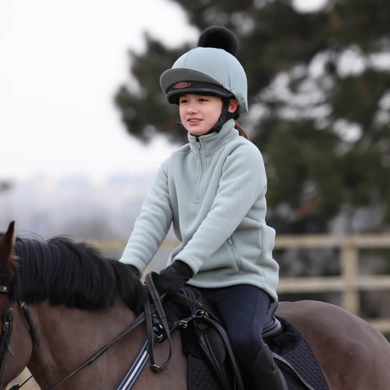 A girl wears the Aubrion React Half Zip Fleece YR Sage while riding a horse outdoors.