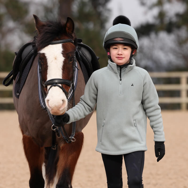 A youth wears the Aubrion React Half Zip Fleece YR Sage, holding a pony’s reins outdoors.