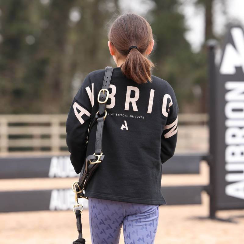 Young rider wears a Shires Aubrion React Sweatshirt Shadow and holds a black lead rein outdoors.