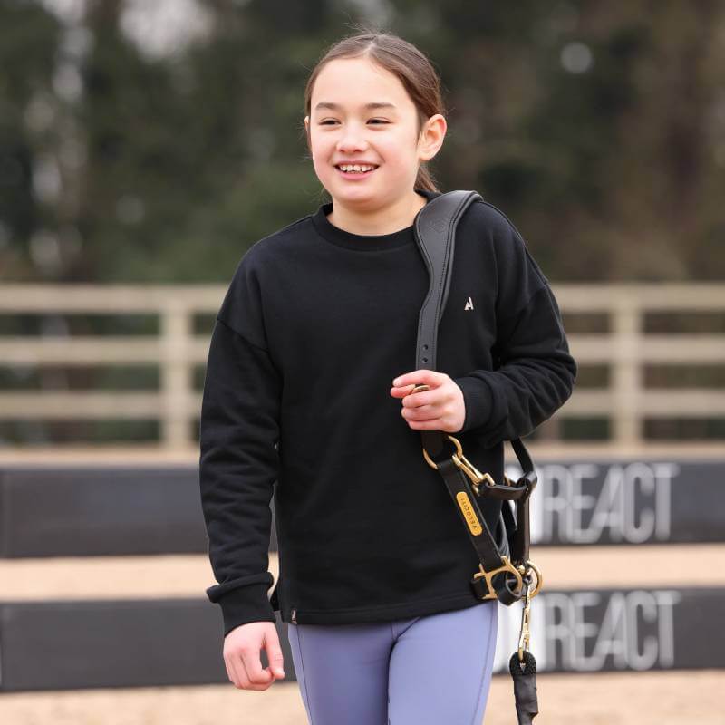 Smiling child in a Shires Aubrion React Sweatshirt Young Rider Shadow holds horse tack outdoors.