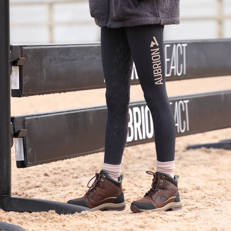 Person in Aubrion React Non-Stop Riding Tights YR Shadow stands by a black equestrian jump.