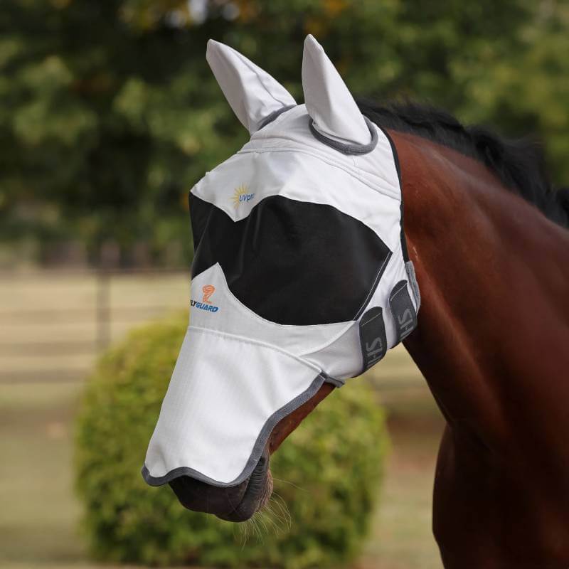 A horse wears the Shires FlyGuard Pro Sun Shade Fly Mask with Ears & Nose outdoors by greenery.