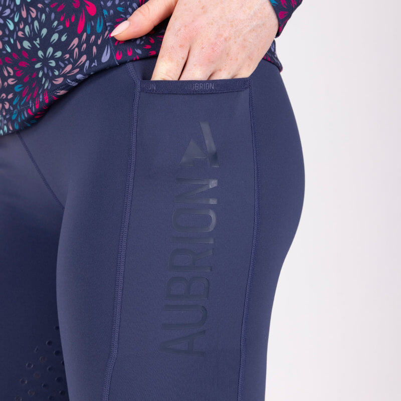 Woman wears Aubrion React Non-Stop Riding Tights Midnight and black boots against plain backdrop.