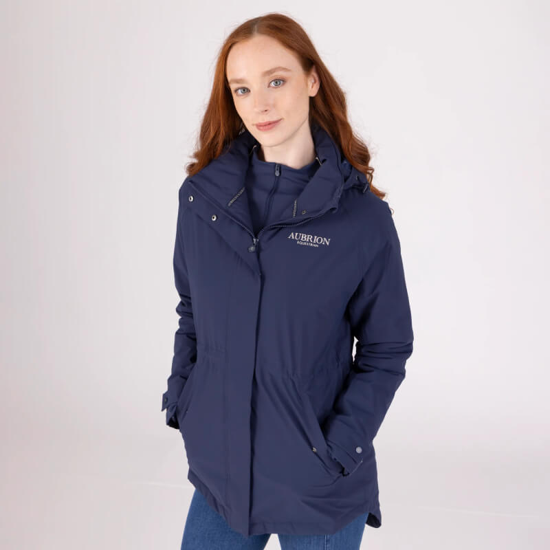 Woman in Aubrion Equestrian Lightweight Coat Midnight, hands in pockets, plain light background.