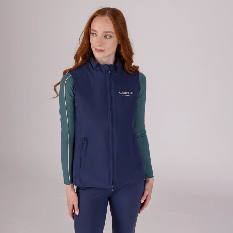 Woman wears Aubrion Equestrian Gilet Midnight and green top, stands against plain backdrop.