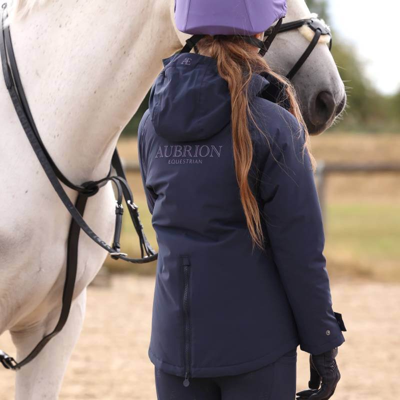 Young rider in Aubrion coat stands by a white horse outdoors, holding the reins.
