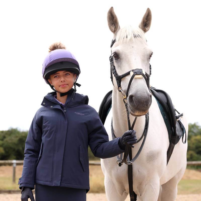 Young rider in helmet, beside white horse, wears Aubrion Equestrian Lightweight Coat Midnight.