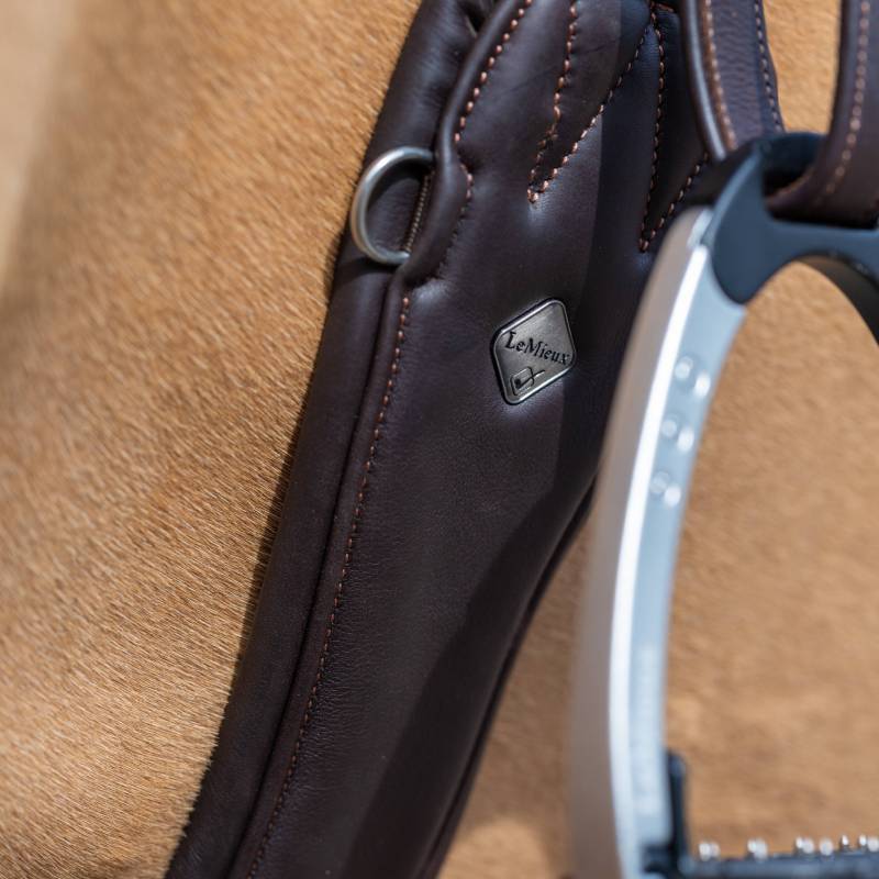 Close-up of a brown saddle with silver stirrup and LeMieux Gel-Tek Anatomic Girth in black.