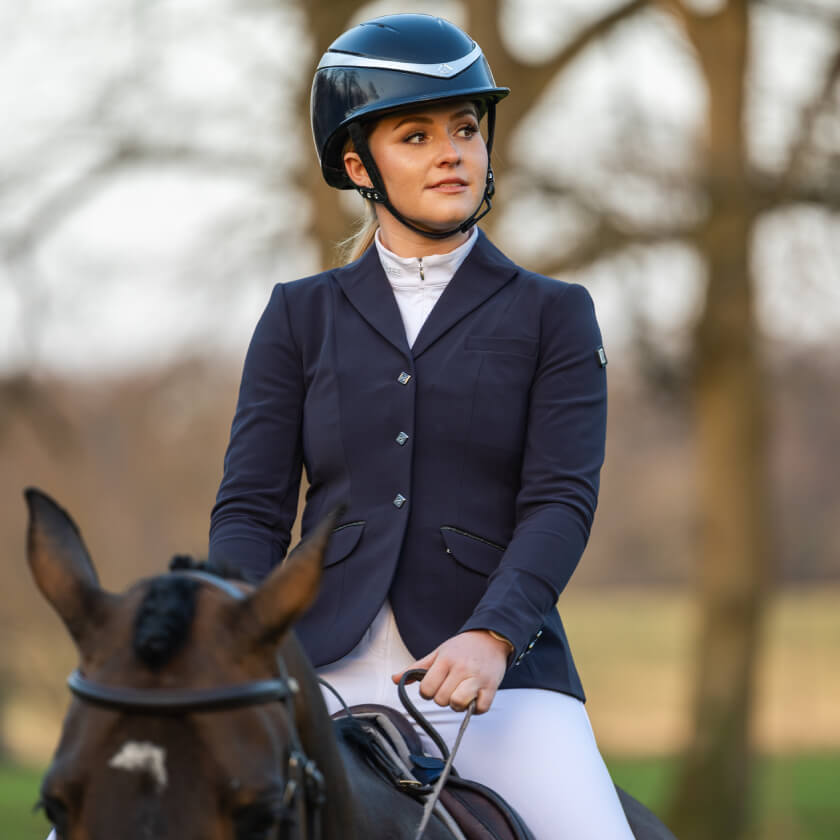 A woman in a LeMieux Dynamique Show Jacket Ladies Navy and helmet sits on a horse outdoors.