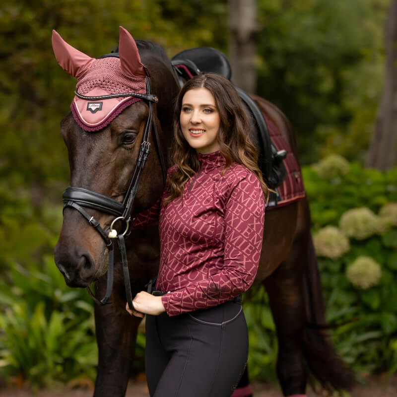 Woman in maroon top stands by horse wearing a LeMieux Loire Fly Hood in Orchid outdoors.