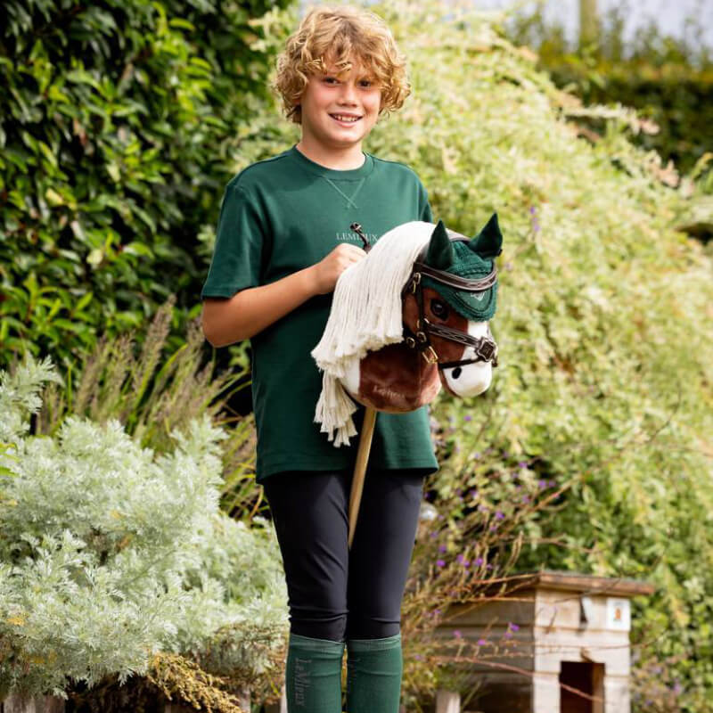Smiling child outdoors with LeMieux Hobby Horse Flash, wearing a green shirt and riding gear.