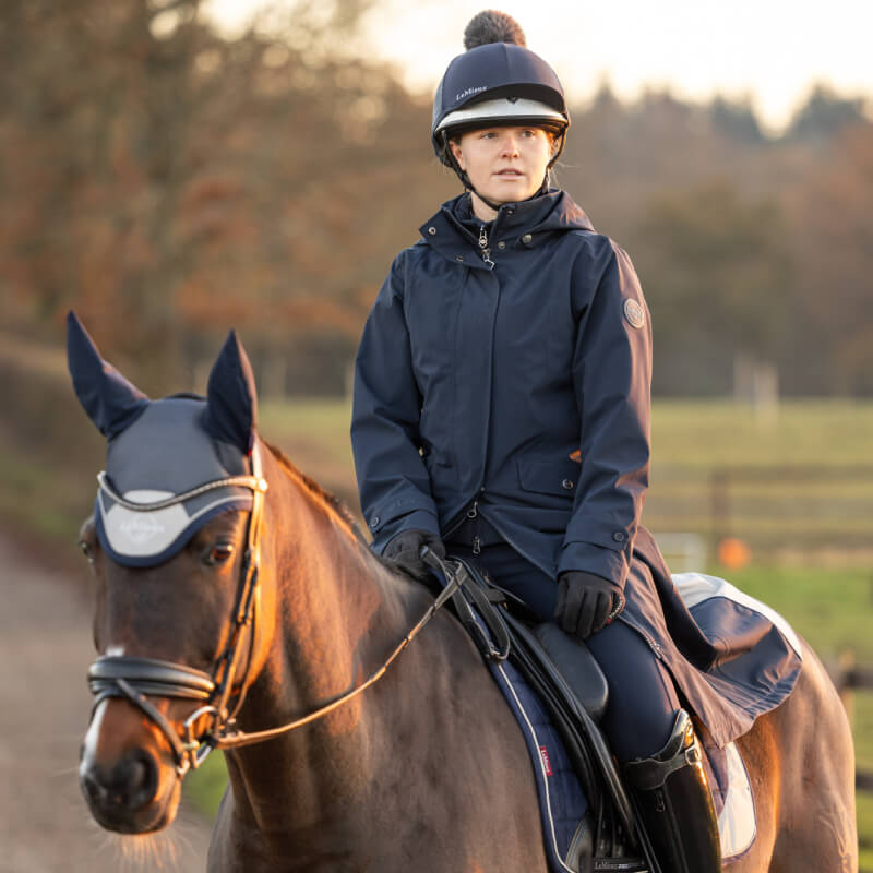 Person in a LeMieux Amelie Lightweight Riding Coat Navy sits on a brown horse outside.