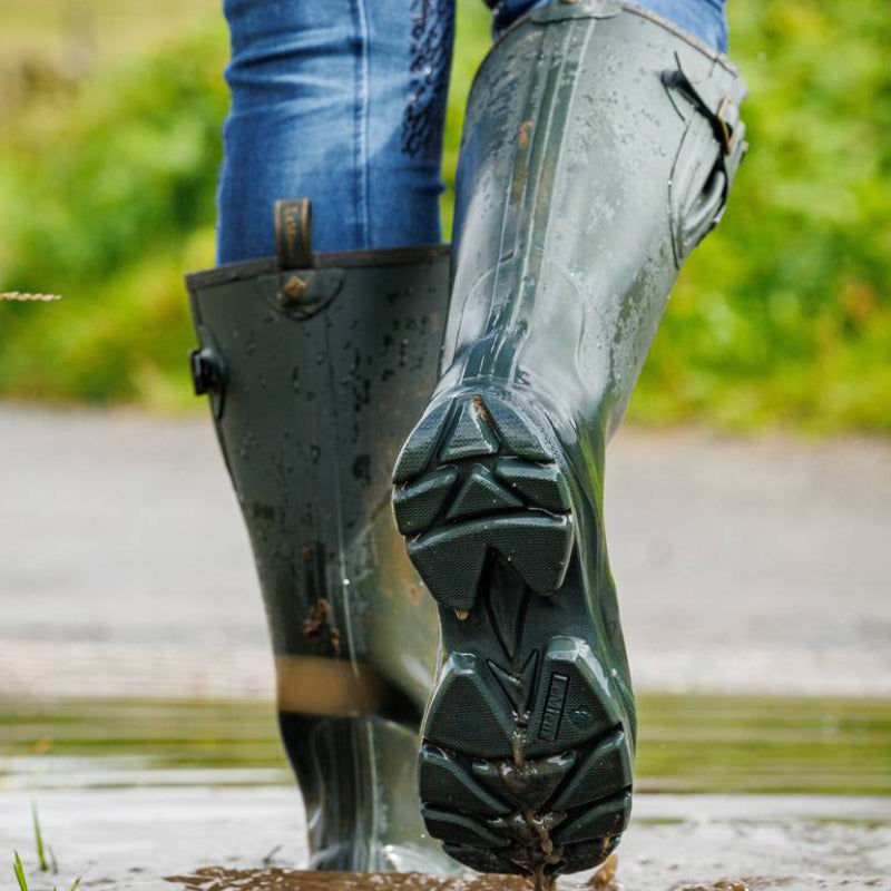 Person wearing LeMieux Stride Wellington Boot Oak standing on grass and rocks outdoors.