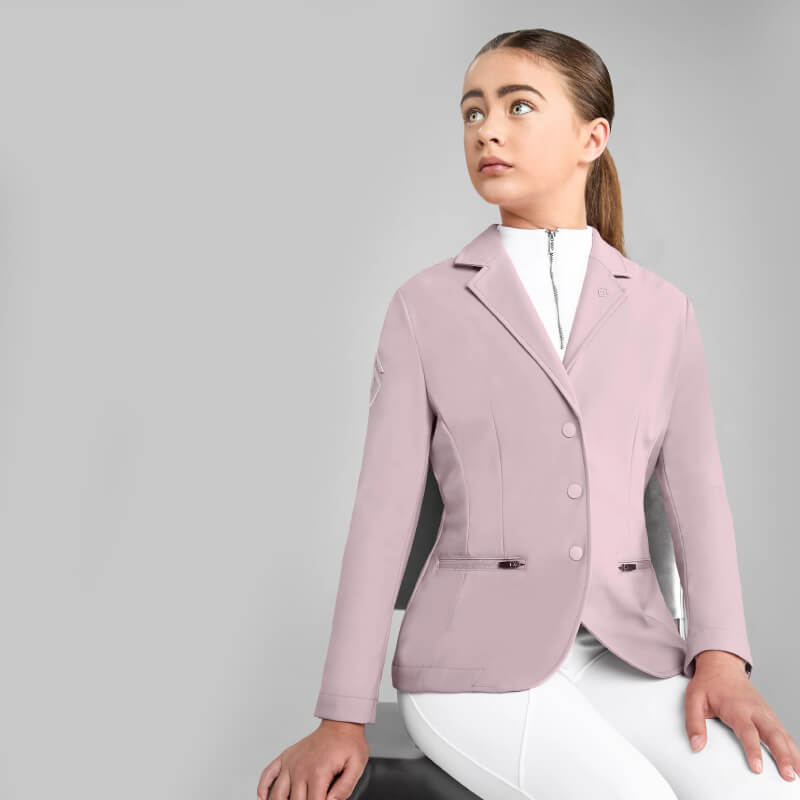 Girl wears Aztec Diamond YR Competition Jacket Pink, white pants; stands by a plain gray background.
