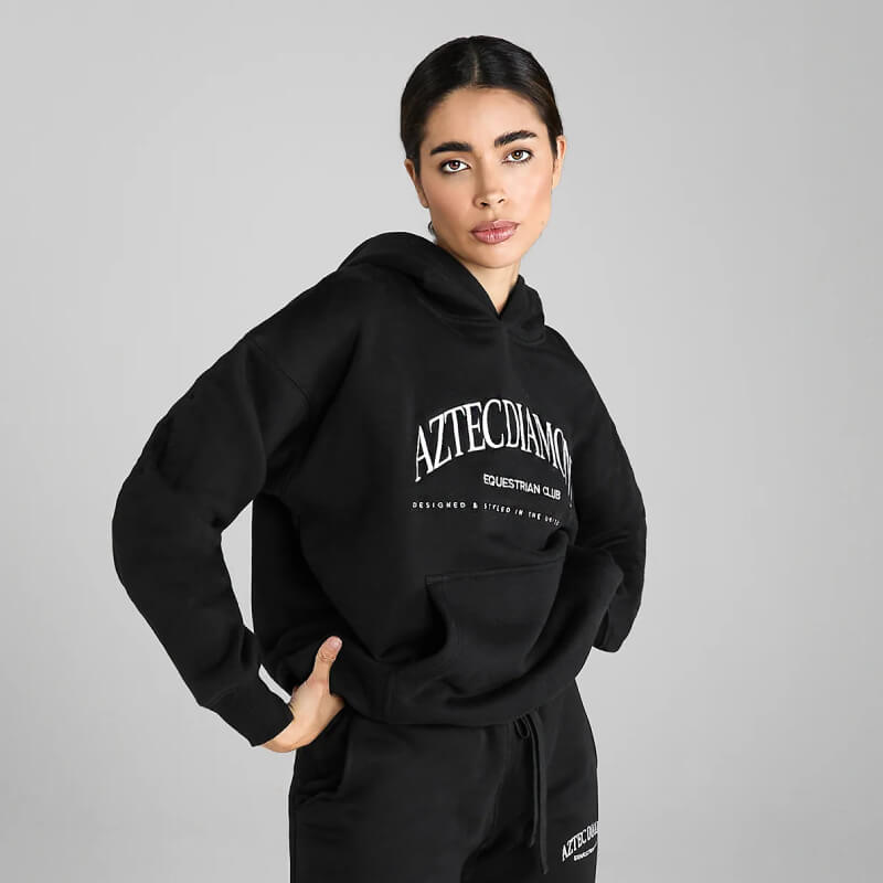 Woman wears Aztec Diamond EQ Club Pull on Hoodie Black, facing the camera.