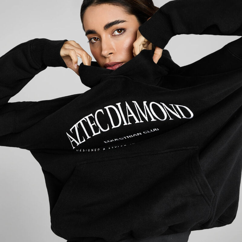 Woman wears Aztec Diamond EQ Club Pull on Hoodie Black, facing the camera.