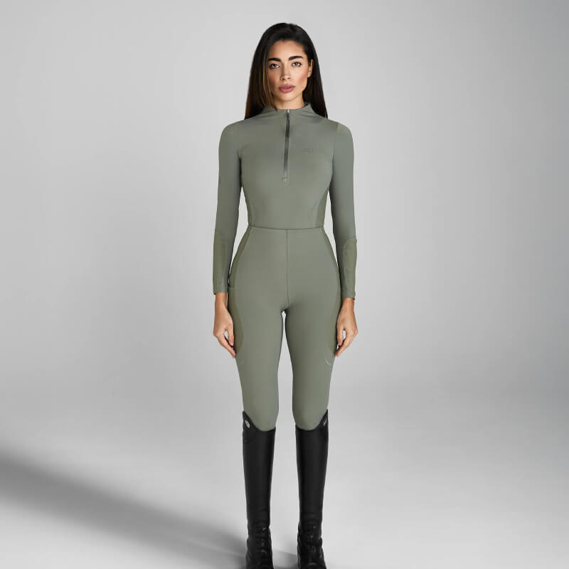 Woman in an Aztec Diamond Define Baselayer Sage jumpsuit and black boots on gray background.