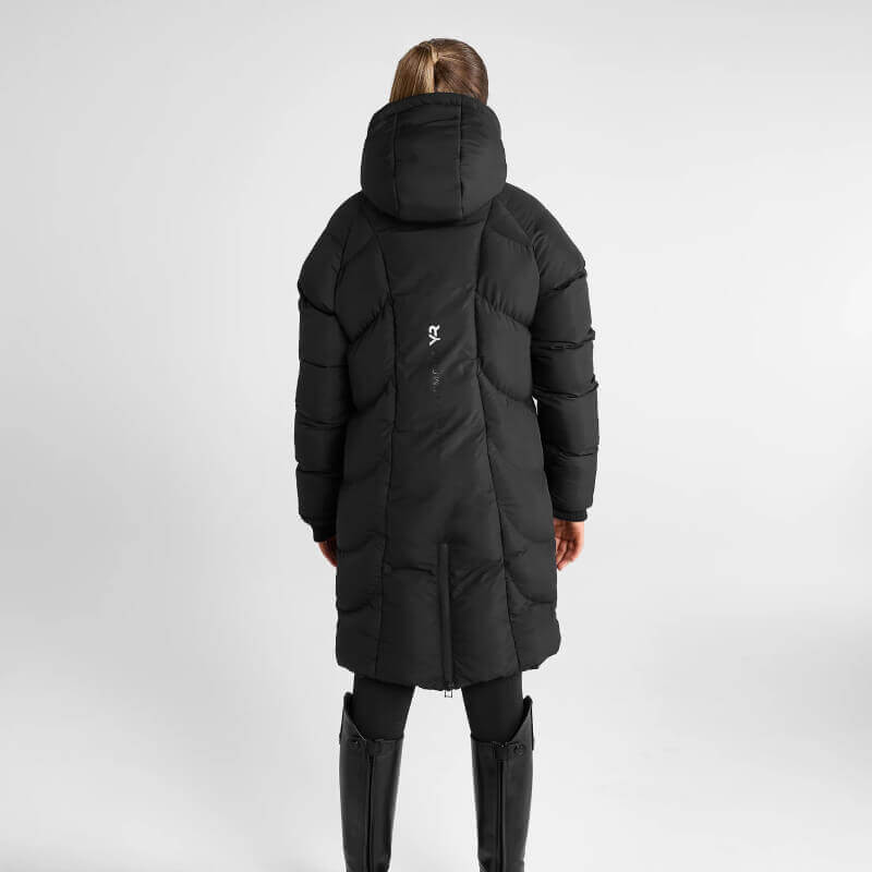Woman wears Aztec Diamond Young Rider Long Puffer Jacket Black, leggings, and boots on gray.
