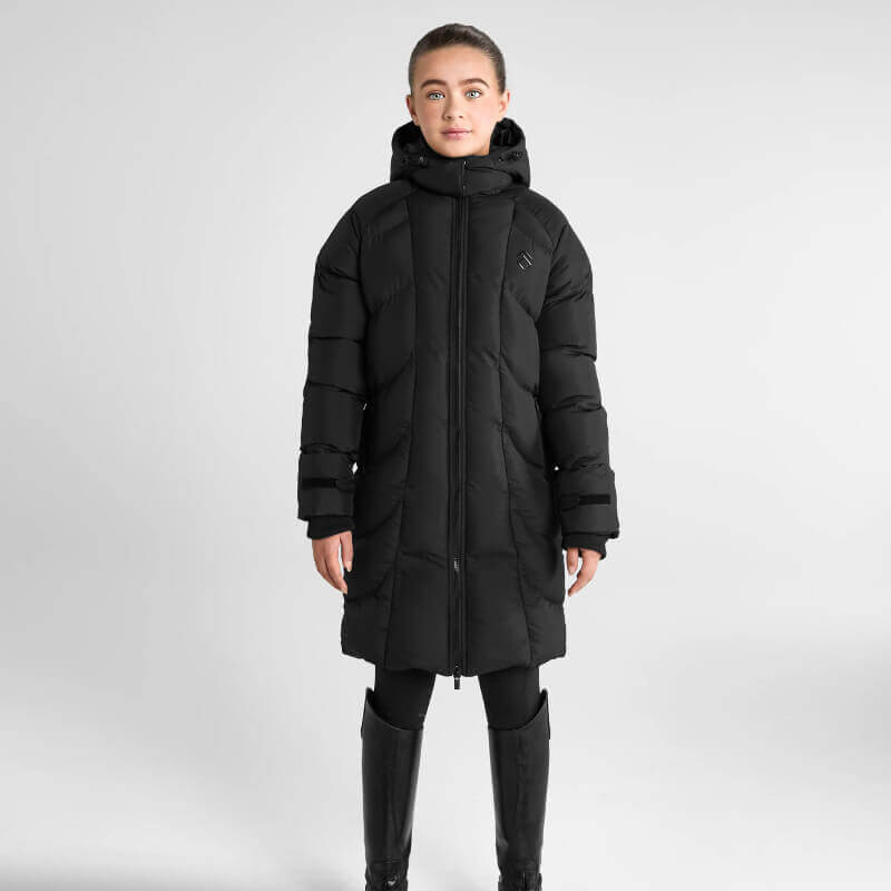 Woman wears Aztec Diamond Young Rider Long Puffer Jacket Black, leggings, and boots on gray.