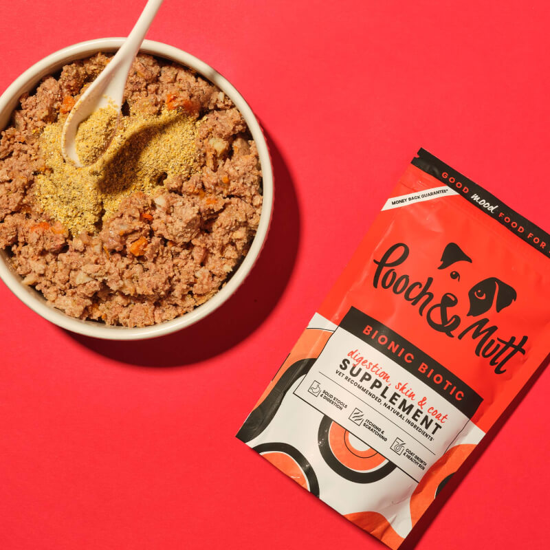 Bowl of dog food and Pooch & Mutt Bionic Biotic 200g supplement on red background.