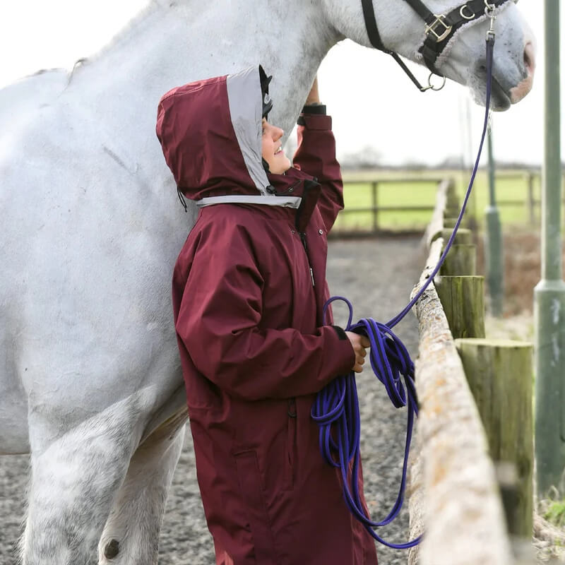 Person in a Reinwear Reincoat Lite Adult Burgundy petting a white horse near a wooden fence.