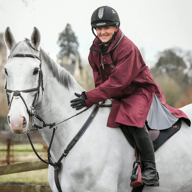 Woman in a Reinwear Reincoat Lite Adult Burgundy and helmet sits on a white horse outdoors.