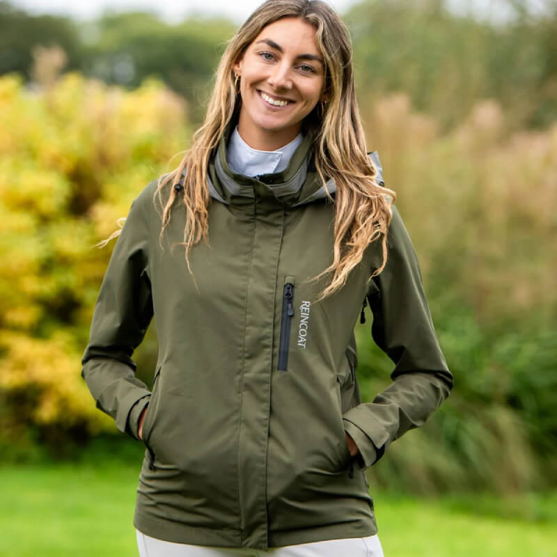 Woman smiles outdoors wearing the Reinwear Reincoat Air Adult Olive, hands in pockets.