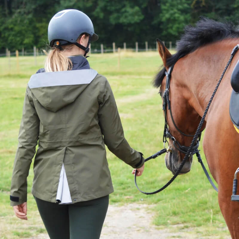 Person in a Reinwear Reincoat Air Adult Olive walks a brown horse on a grassy field.