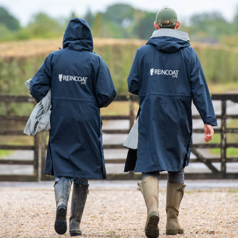 Two people in Reinwear Reincoat Lite Adult Midnight Navy walk on a gravel path near a wooden fence.