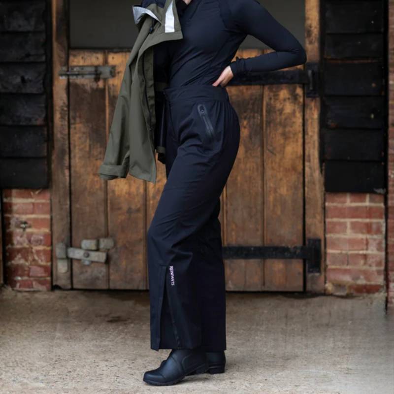 Person in Reinwear Reinpants Lite Black and boots stands by a rustic door, holding a green jacket.