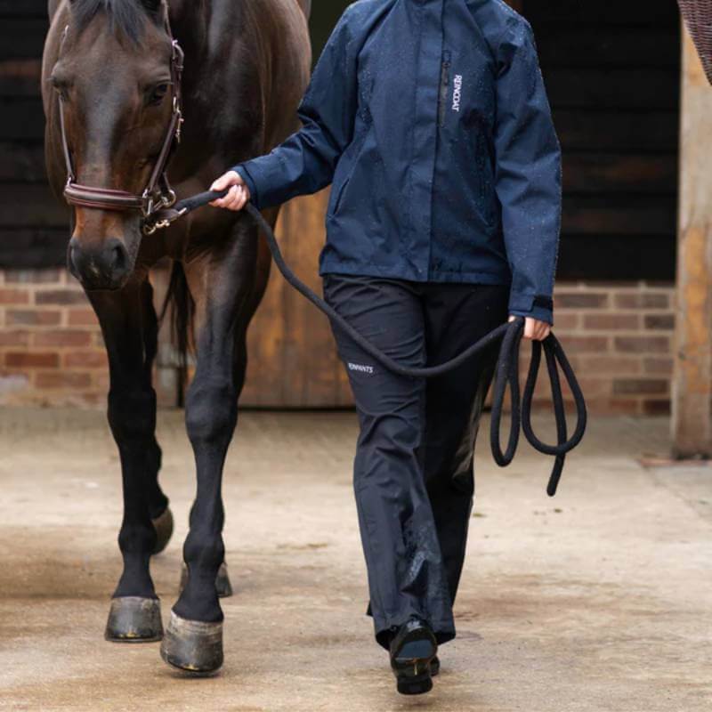 Person in a navy rain jacket and Reinwear Reinpants Lite Black walks a brown horse near a stable.