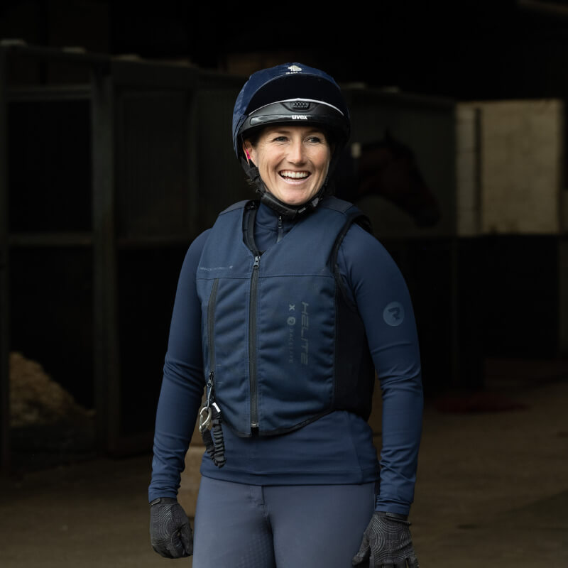 Smiling woman in Racesafe Motion Air Jacket Navy Adult stands near the stables.