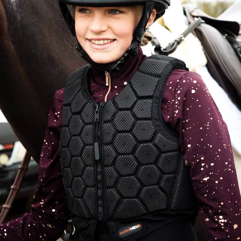Young rider in a helmet and Evoke HEX6 Body Protector stands next to a horse.