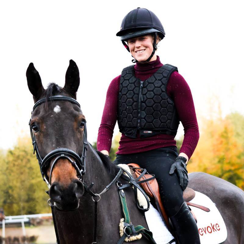 Rider in Evoke HEX6 Body Protector (Black) smiles on a dark brown horse outdoors.