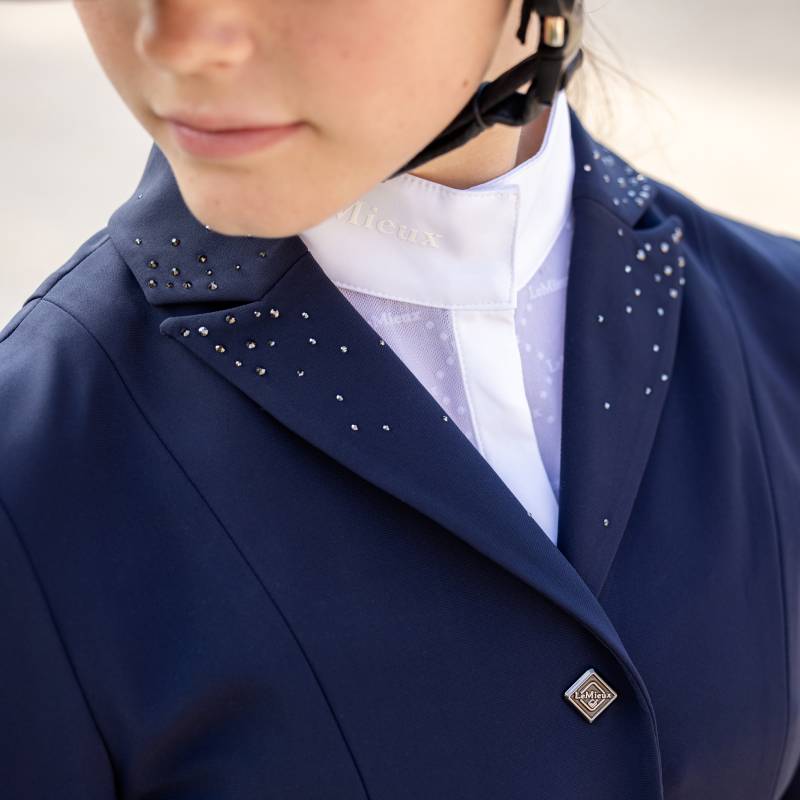 Wearing a LeMieux Junior Pro Show Jacket Navy, a rider stands outdoors among greenery and flowers.