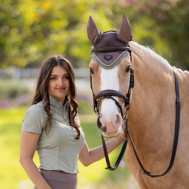 A white horse wears a LeMieux Loire Fly Hood Walnut and riding gear on a dirt path.