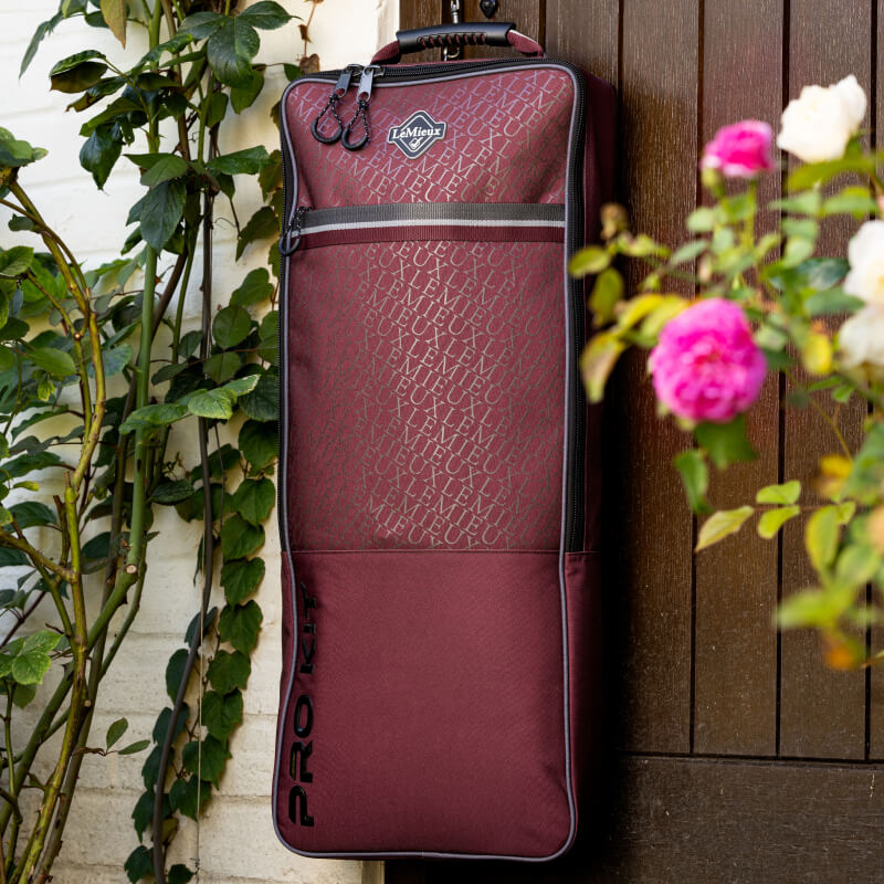 A LeMieux Elite Pro Bridle Bag Burgundy hangs on a brown door near pink roses.