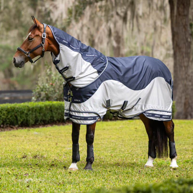 A brown horse in a LeMieux Arika Shower-Tek Fly Rug Navy grazes in a fenced grassy area.