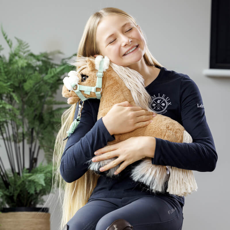 Smiling girl hugs a LeMieux Toy Pony Cloudy indoors, plant in the background.