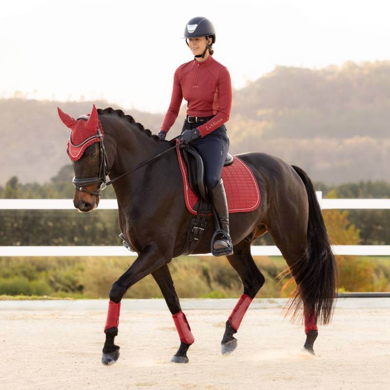 A rider in red on a dark horse wears a LeMieux Suede Dressage Square Russet in an outdoor arena.