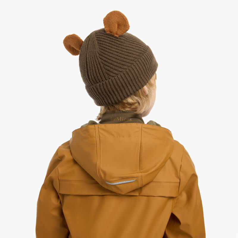 Smiling child in a LeMieux Mini Bear Beanie Alpine and brown jacket, on a white background.
