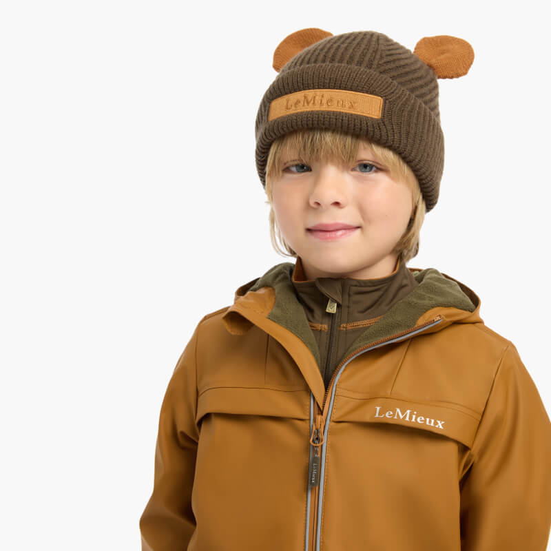 Smiling child in a LeMieux Mini Bear Beanie Alpine and brown jacket, on a white background.