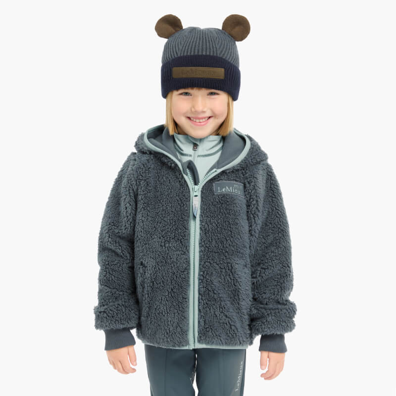 Smiling child in fuzzy gray jacket wears LeMieux Mini Bear Beanie Petrol against plain background.