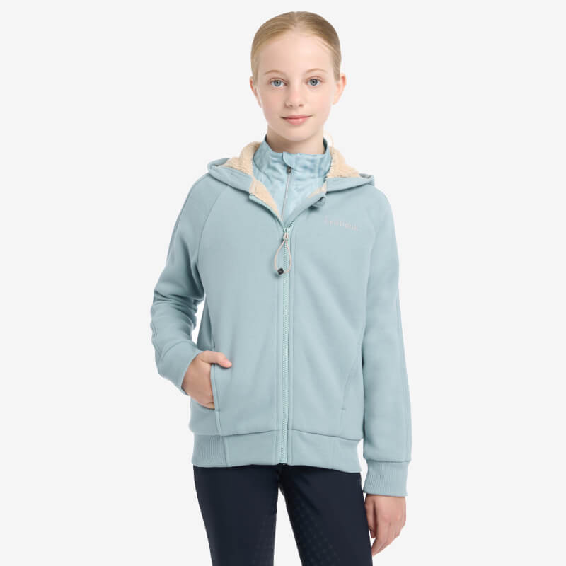 Young girl in a LeMieux Young Rider Hollie Lined Hoodie Glacier, hands in pockets, on white.