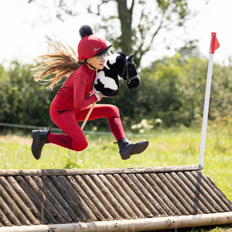 Girl in red jumps over a wooden obstacle riding the LeMieux Hobby Horse Razzle outdoors.