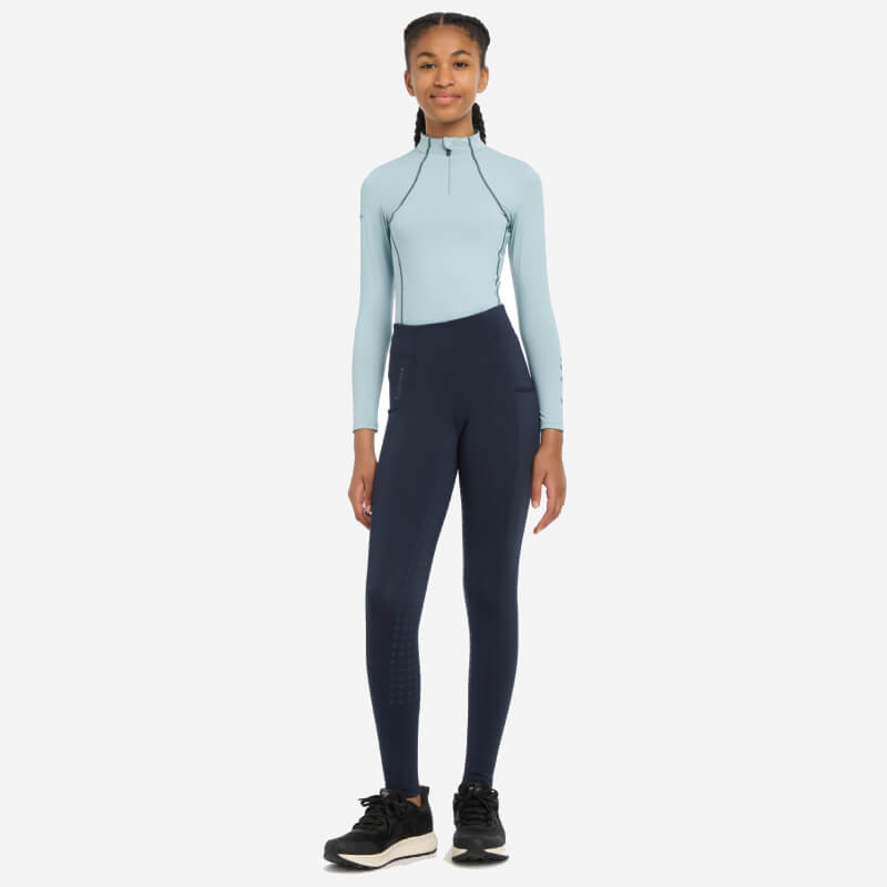 A woman in LeMieux Young Rider Orla Pull Ons Navy and a light blue top stands smiling forward.